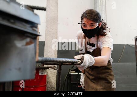 Seriöse Schmiede in Schutzmaske und Handschuhe in der Werkstatt Metallarbeiten Stockfoto