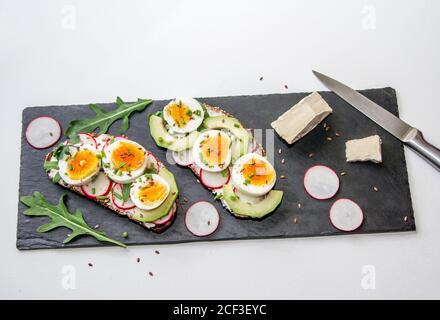 Sandwiches mit weich gekochtem Ei, Avocado, Rettich, Rucola, grünen Zwiebeln und Leinsamen auf einem schwarzen Steinteller. Gesunde Snacks. Stockfoto