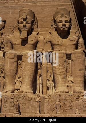Zwei der vier sitzenden Kolosse, jeweils 20 m (66 ft) hoch, des lang herrschenden façade. Dynastie New Kingdom Pharao, Ramesses II, die die   des Großen Tempels dominieren, den er im 13. Jahrhundert v. Chr. in Abu Simbel, Assuan Governate, Ägypten, errichtete. Kleinere Bildnis seiner Frauen und Kinder stehen neben den Kolossen und zwischen ihren Füßen. Die zwei massiven Felstempel bei Abu Simbel stehen am westlichen Ufer des Nasser Sees in Nubia, Oberägypten. Sie wurden 1968 auf einen künstlichen Hügel hoch über dem Assuan-Staudamm verlegt, um zu verhindern, dass sie vom aufsteigenden Wasser des Sees umspült werden. Stockfoto