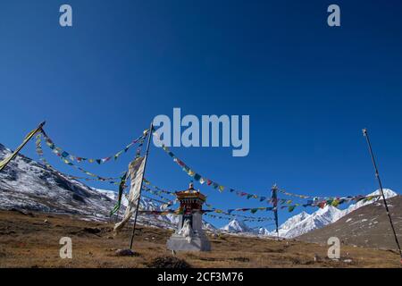 Ein buddhistischer Schrein, umgeben von bunten Gebetsfahnen an einem windigen, klaren Tag mit einer Reihe von Schneespitzen im Hintergrund und blauem Himmel. Stockfoto