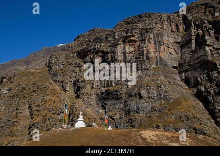 Ein buddhistischer Schrein, umgeben von bunten Gebetsfahnen an einem windigen, klaren Tag mit einer Reihe von Schneespitzen im Hintergrund und blauem Himmel. Stockfoto