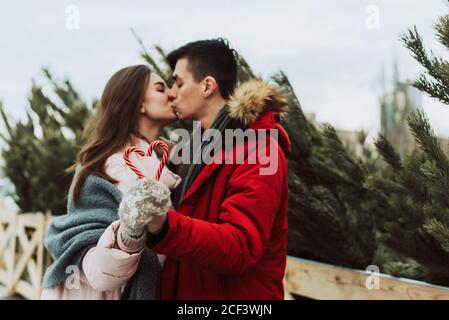 Eine junge Frau und ein Mann halten herzförmige Karamellstöcke und küssen vor dem Hintergrund des Weihnachtsbaum-Basars in der Stadt. Stockfoto