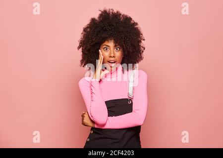 Erstaunt afroamerikanische Frau trägt rosa Rollkragen und schwarz insgesamt Das Gesicht berühren und die Kamera mit offenem Mund betrachten überrascht Mit Nachrichten Stockfoto