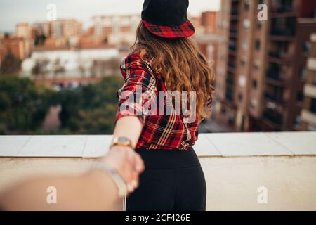 Rückansicht einer jungen Frau im trendigen Outfit, die die Hand eines anonymen Freundes auf dem Balkon vor dem verschwommenen Hintergrund der modernen Stadt hält Stockfoto