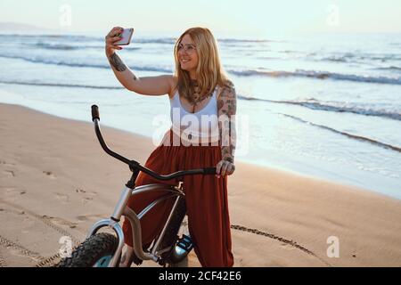 Lächelnde, tätowierte Frau in einer Brille, die auf dem Smartphone fotografiert, während sie mit dem Fahrrad am ruhigen Strand steht Stockfoto