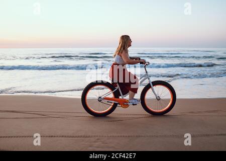 Seitenansicht der molligen modernen glücklichen Frau in hellem Rock, die in der Dämmerung Fahrrad entlang der malerischen Küste des Ozeans reitet Stockfoto