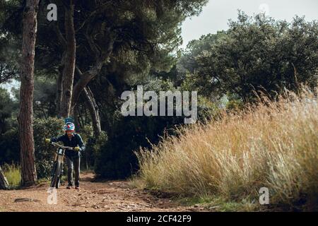 Hübscher Mann im Helm, der beim Tragen auf die Kamera schaut Mountainbike-Bike nach dem Training auf der Wiese Stockfoto