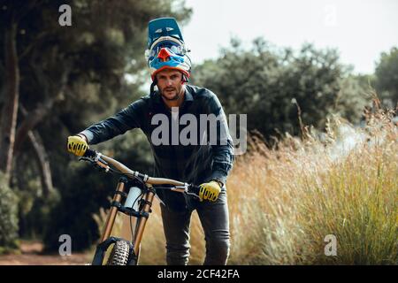 Hübscher Mann im Helm, der beim Tragen auf die Kamera schaut Mountainbike-Bike nach dem Training auf der Wiese Stockfoto