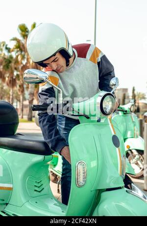 Junger gutaussehender Mann im farbigen Kapuzenpulli und Helm konzentriert stehend Beim Starten des grünen Rollers Stockfoto
