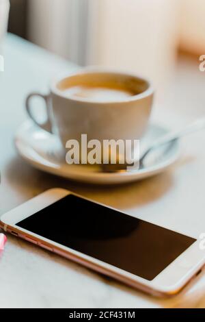Weißes Smartphone und Tasse mit Kaffee auf Marmortisch in Cafe mit kleinem Notizblock und stylischer Sonnenbrille Stockfoto