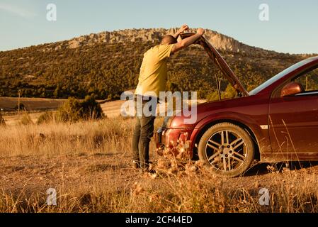 Seitenansicht des nicht erkennbaren erwachsenen Mannes, der die Kapuze öffnet Motor eines kaputten Autos nach einem Unfall auf dem Land Stockfoto