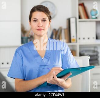 Positive Brünette Frau Arzt im Büro mit Zwischenablage in den Händen, Schreiben Krankengeschichte Blatt Stockfoto