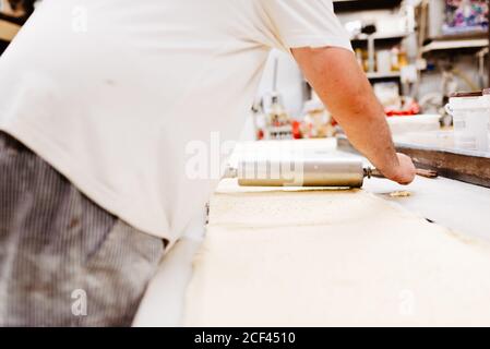 Anonymer übergewichtiger Mann in Uniform rollt frischen weichen Teig auf Tisch beim Kochen Gebäck in der Bäckerei Stockfoto