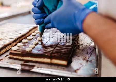 Nahaufnahme anonymer Mitarbeiter in Handschuhen quetschen Creme auf Frische Schokoladenkuchen auf Tablett in der Bäckerei Stockfoto