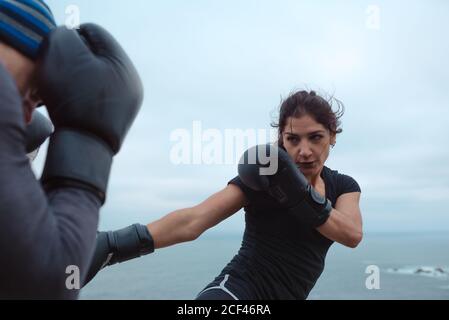 Seitenansicht von Mann und Frau in Boxhandschuhen, die sich gegenseitig schlagen, während sie auf einer Klippe gegen das Meer und den wolkenlosen Himmel stehen Stockfoto