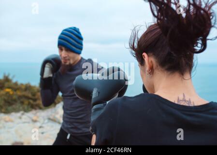 Seitenansicht von Mann und Frau in Boxhandschuhen, die sich gegenseitig schlagen, während sie auf einer Klippe gegen das Meer und den wolkenlosen Himmel stehen Stockfoto