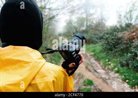 Rückansicht von nicht erkennbaren weiblichen Touristen in lebhaft gelben Regenmantel Und schwarze Kapuze mit Digitalkamera, die alleine auf der Straße steht Zwischen Nebelwald Stockfoto