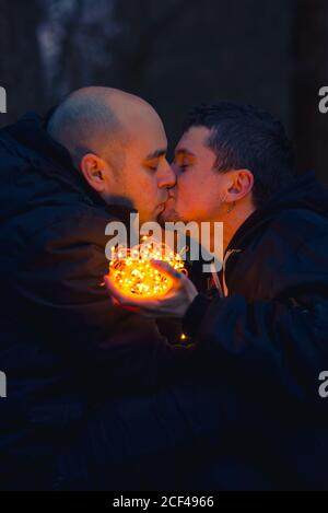 Seitenansicht des homosexuellen Paares küssen mit beleuchteten Lichterketten Sitzen im Murk Wald mit Hund am Abend Stockfoto