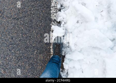 Draufsicht auf Ernte unkenntlich Person in Jeans und Trekking Schuhe stehen auf Asphalt verschneiten Straße am Tag Stockfoto