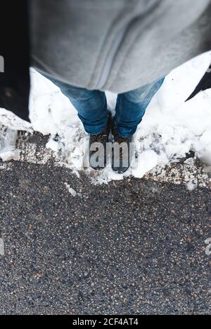 Draufsicht auf Ernte unkenntlich Person in Jeans und Trekking Schuhe stehen auf Asphalt verschneiten Straße am Tag Stockfoto