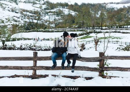 Romantischer Mann und Frau in Winterkleidung küssen, während sie bei schneebedecktem Wetter auf einem kleinen Holzzaun auf der Natur sitzen Stockfoto