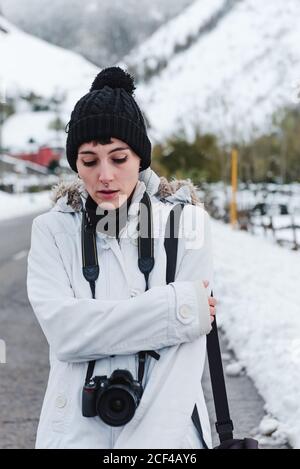 Frau in weißer Winterjacke mit Kapuze und schwarzer Hose Wandern inmitten der Asphaltstraße zwischen verschneiten Bergen mit Kamera Am Hals Stockfoto