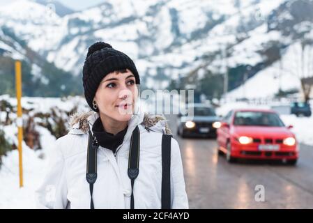 Frau in weißer Winterjacke mit Kapuze und schwarzer Hose Wandern inmitten der Asphaltstraße zwischen verschneiten Bergen mit Autos Im Hintergrund Stockfoto