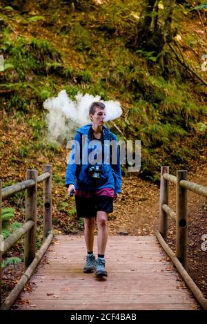 Frau mit Kamera, die auf einer Holzbrücke hinter dichtem Rauch steht Beim Rauchen stumpf während Camping im Wald Stockfoto