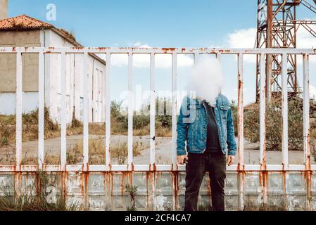 Unkenntlich männlich im lässigen Outfit Ausatmen vape Wolke vor Von Gesicht steht gegen rostigen Zaun des verlassenen Industriegebäude Stockfoto