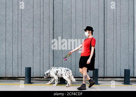 Trendy nachdenkliche Frau in Gesichtsmaske zu Fuß mit Hund in der Stadt Stockfoto