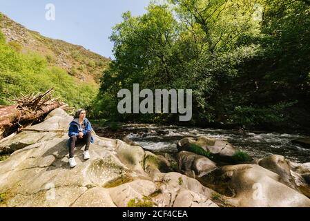 Ruhige Touristen in Oberbekleidung sitzen auf felsigen Ufer und bewundern Tolle Aussicht auf den schnellen Gebirgsfluss beim Entspannen während des Trekkings In Hoces del Esva Stockfoto