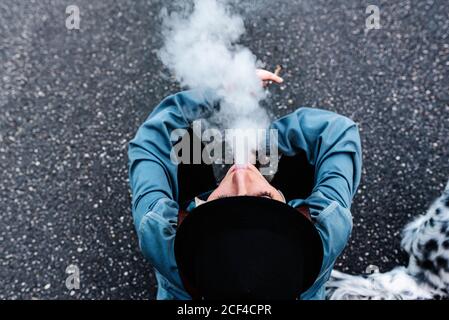 Von oben Blick auf nicht erkennbare weibliche in stilvollen schwarzen Hut Und legere Kleidung sitzt auf Asphalt Straße und Rauchen Zigarette Während des Spaziergangs ruhen Stockfoto