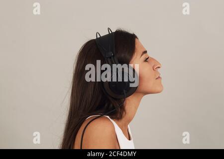 Seitenansicht der nachdenklichen jungen attraktiven dunkelhaarigen weiblichen tragen Stereo-Kopfhörer und Musikhören auf grauem Hintergrund in studio Stockfoto