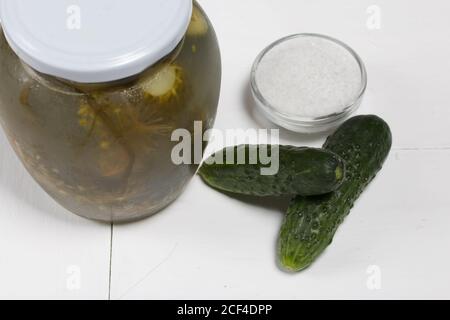 Glas mit eingelegten Gurken. In der Nähe sind frische Gurken und ein Behälter mit Salz. Auf einer lackierten Plattenoberfläche. Stockfoto