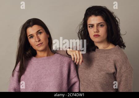 Zufriedene junge charmante dunkelhaarige Frauen tragen Pullover und suchen Bei der Kamera, während sie auf grauem Hintergrund im Studio zusammen stehen Stockfoto