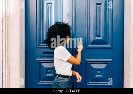 Hübsche ethnische Frau in weißem T-Shirt und Jeans klopft an blaue Tür und schaut weg Stockfoto