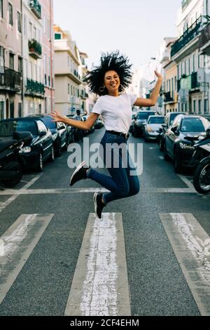Junge, fröhliche afroamerikanische Frau in Jeans und weißem T-Shirt, die tagsüber vor Freude auf der Straße der Stadt hüpft Stockfoto