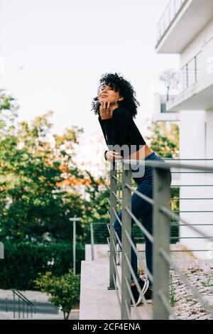 Von unten junge schöne Frau mit fliegenden lockigen Haaren, die auf dem Balkon steht, sich auf das Geländer lehnt und wegschaut Stockfoto