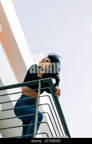 Von unten junge schöne Frau mit fliegenden lockigen Haaren, die auf dem Balkon steht, sich auf das Geländer lehnt und zurückblickt Stockfoto