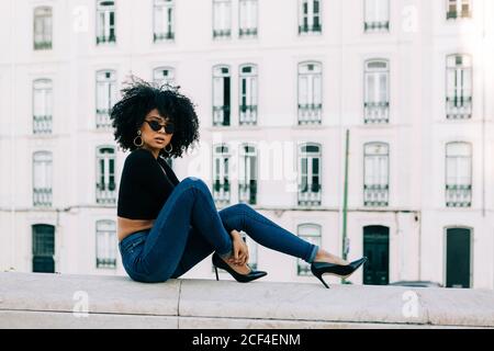 Junge trendige afroamerikanische Frau in Jeans und Crop Top sitzt auf Steinbrüstchen und schaut über Sonnenbrillen Stockfoto
