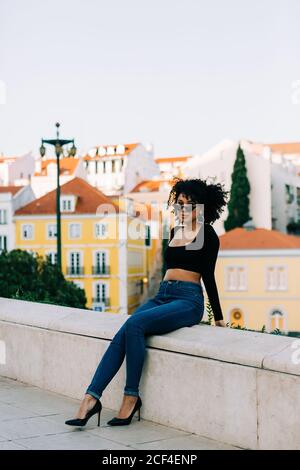 Junge trendige afroamerikanische Frau in Jeans und Crop Top sitzt auf Steinbrüstchen und schaut über Sonnenbrillen Stockfoto