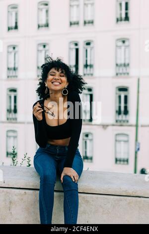 Junge trendige afroamerikanische Frau in Jeans und Crop Top sitzen auf Steinbrüstchen und schauen in die Kamera Stockfoto