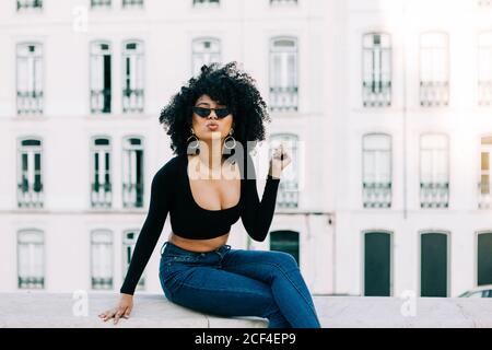 Junge trendige afroamerikanische Frau in Jeans und Crop Top sitzt auf Steinbrüstchen und schaut über Sonnenbrillen Stockfoto