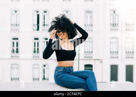 Junge trendige afroamerikanische Frau in Jeans und Crop Top sitzt auf Steinbrüstchen und schaut über Sonnenbrillen Stockfoto