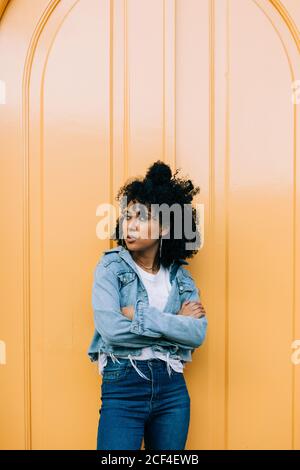 Junge afroamerikanische Frau in Jeans und Denim-Jacke, die sich an der gelben Tür lehnt und wegschaut Stockfoto