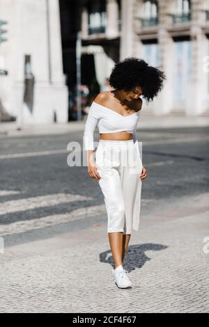 Schöne afroamerikanische Frau in weißem modischem Outfit, die vor städtischem Hintergrund am Straßenrand steht Stockfoto