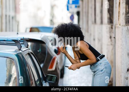 Seitenansicht einer jungen ethnischen Frau, die auf das Autofenster schaut und sich schminkt, während sie auf einem städtischen Hintergrund steht Stockfoto