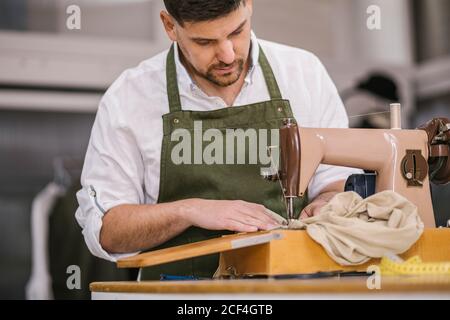 Diligent fokussierten männlichen Schneider in Schürze nähen Outfit Details mit Moderne Nähmaschine am Tisch, während die Schaffung exklusiver Kleidung Sammlung In zeitgenössischem Arbeitsstudio Stockfoto