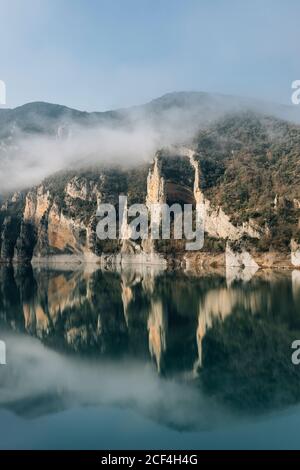 Herrliche Landschaft von ruhigen See mit spiegelnden Wasseroberfläche umgeben Durch raue felsige Berge von Montsec Bereich mit dichten bedeckt Nebel an kalten Tagen in Spanien Stockfoto