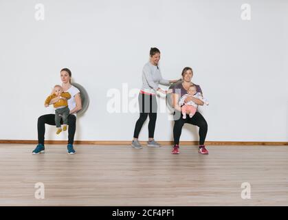 Freundliche weibliche Fitness-Ausbilderin hilft Frauen mit Babys zu führen Kniebeugen in der Nähe der Wand während des Trainings im Fitnessstudio Stockfoto